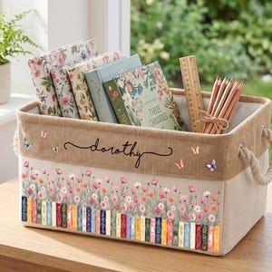 Puede incluir: Una cesta de almacenamiento rectangular de tela con asas de cuerda, con un diseño estampado de flores silvestres coloridas y una fila de libros. El nombre "dorothy" está impreso en letra cursiva negra en la banda superior, junto a pequeñas mariposas. Contiene libros y una regla de madera.