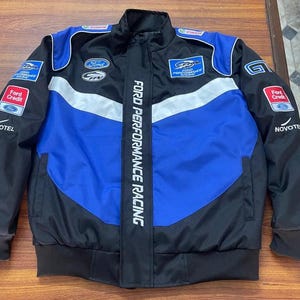 Pode incluir: Um casaco de corrida preto, azul e branco com o texto FORD PERFORMANCE RACING na vertical ao longo do fecho central. O casaco inclui vários logótipos de patrocinadores como Ford, Ford Credit, Castrol e Novotel no peito e mangas.