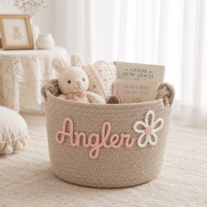 Puede incluir: Una cesta de almacenamiento de cuerda trenzada beige con el nombre Angler en letras cursivas rosas y una aplicación de flor blanca. La cesta contiene un peluche de conejo color crema y dos libros titulados Guess How Much I Love You y Bunny Roo, I Love You.