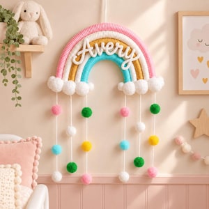 Könnte beinhalten: Ein Wandbehang in Regenbogenform aus umwickeltem Garn in Pink, Gelb und Türkis mit weißen Wolken an der Basis. Der Name Avery ist in weißer Schreibschrift in der Mitte angebracht. Vier weiße Schnüre hängen herab, die mit bunten Pompons verziert sind.