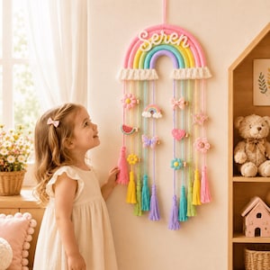 May include: A wall-hanging rainbow organizer featuring the name Seren in white script. The rainbow has pink, yellow, green, and purple arches with cream-colored fringe. Hanging from it are colorful yarn strands with clips holding felt shapes.