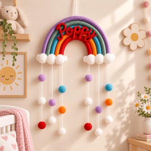 Könnte beinhalten: Ein Wandbehang in Regenbogenform aus umwickeltem Garn in Lila, Blau, Türkis, Orange und Rot, mit dem Namen Poppy in roter Schrift. Weiße Pompon-Wolken bilden die Basis, von denen weiße Schnüre mit bunten Pompons nach unten hängen.