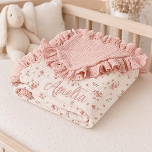 May include: A folded muslin baby blanket featuring a cream base with a delicate pink floral pattern and a solid pink ruffled border. The name Amelia is embroidered in cursive script on the front. The fabric has a textured, crinkled appearance.