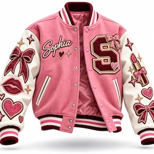May include: A pink varsity jacket with white sleeves, featuring a maroon letter S patch, the name Sophia in script, and various glittery patches including bows, hearts, lips, and a lipstick tube. The jacket has striped ribbed trim and snap buttons.