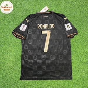 Może przedstawiać: Czarna koszulka piłkarska z nazwiskiem RONALDO i numerem 7 w złotym kolorze na plecach. Materiał posiada tonalny, powtarzający się wzór godła narodowego Portugalii. Złote logo Puma znajdują się na ramionach, a na rękawach widoczne są naszywki.