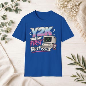 Puede incluir: Una camiseta azul real de manga corta con un estampado gráfico de una computadora antigua con una cara triste en la pantalla. El texto dice Y2K WAS MY FIRST TRUST ISSUE en una mezcla de colores azul, rosa y blanco con un icono de reloj de arena.
