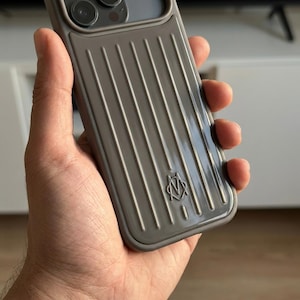 May include: A taupe-colored smartphone case featuring a series of vertical, raised ridges across the back panel. The design includes a small, embossed logo near the bottom center, consisting of the letters M and V intertwined within a circular frame.
