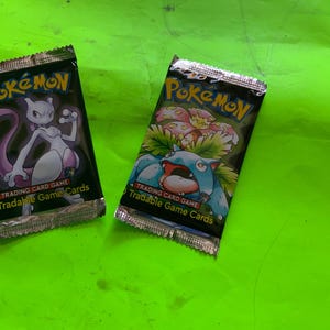 Op de afbeelding: Twee verzegelde Pokémon trading card game booster packs. Het linkerpakje bevat een illustratie van het personage Mewtwo in paars en wit, terwijl het rechterpakje een blauw-groene Venusaur toont. Beide pakjes hebben metallic zilveren randen.