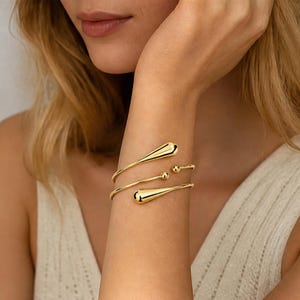 Può includere: Un bracciale a fascia in metallo color oro con design avvolgente. Il bracciale è composto da un filo sottile e lucido che si avvolge attorno al polso, terminando con due terminali a forma di goccia e due piccoli accenti sferici.