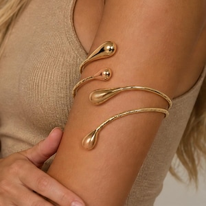 May include: A gold-toned metal arm cuff featuring a spiral design that wraps around the upper arm. The piece is constructed from a continuous wire that terminates at both ends in smooth, polished, teardrop-shaped accents with a reflective finish.
