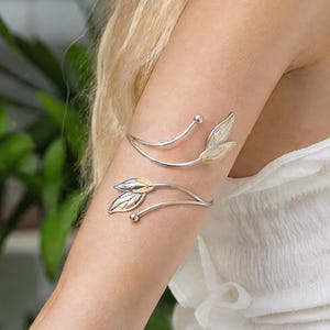 Peut inclure: Un bracelet de bras en métal argenté composé de deux bandes incurvées qui s'enroulent autour du bras. Chaque bande se termine par une petite perle sphérique à une extrémité et deux ornements en forme de feuille polie avec des détails texturés.