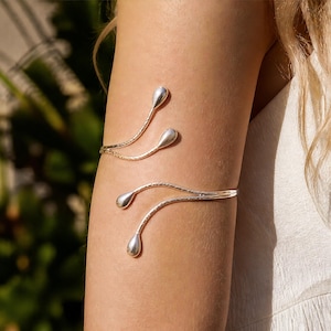 May include: A silver-toned arm cuff featuring a bypass design with four distinct, smooth, teardrop-shaped terminals. The two main bands of the cuff have a subtle, textured, hammered finish that catches the light, creating a delicate and organic appearance on the upper arm.