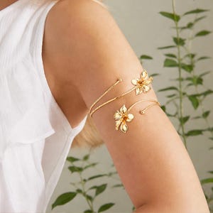 Può includere: Un bracciale da braccio in metallo color oro con un design a spirale che avvolge la parte superiore del braccio. Il pezzo è decorato con due dettagliati motivi floreali a cinque petali su ciascuna estremità, con piccole sfere alle estremità.