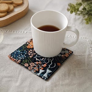 Puede incluir: Una taza de cerámica blanca con estrías verticales llena de un líquido marrón oscuro, apoyada sobre un posavasos de tela cuadrado con fondo azul marino oscuro y un intrincado bordado floral multicolor en tonos naranja, azul y blanco.