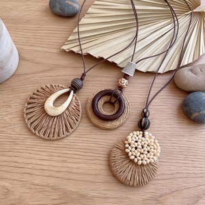 Puede incluir: Tres collares con cordones marrón oscuro y colgantes circulares. Un colgante tiene una forma de lágrima crema sobre rafia tejida, otro un anillo de madera marrón oscuro sobre rafia tejida y otro cuentas crema sobre rafia tejida.
