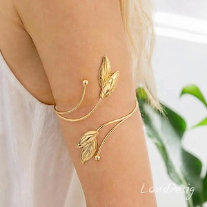 Può includere: Un bracciale da braccio in metallo color oro dal design aperto con due rami curvi. Ogni ramo termina con una coppia di motivi a foglia stilizzati e lucidi e una piccola perla sferica all'estremità di un filo sottile.
