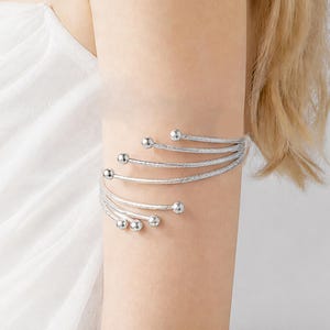 May include: A silver-toned arm cuff featuring a multi-strand spiral design. The accessory consists of several curved metal wires that wrap around the arm, each ending in a small, smooth spherical bead. The surface of the wires has a textured finish.