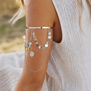 Op de afbeelding: Een zilverkleurige metalen bovenarmband met een gladde, stevige band die om de bovenarm sluit. Aan de band hangen meerdere fijne zilveren kettinkjes in lagen, versierd met talloze kleine, ronde en reflecterende metalen schijfjes.