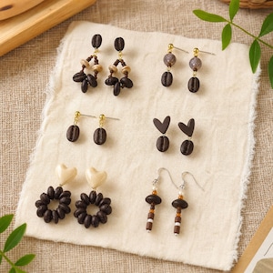 May include: Six pairs of earrings featuring coffee bean motifs, brown wooden beads, cream-colored heart shapes, and metallic hardware. The collection includes dangling designs with various bead combinations, stud-style coffee beans, and heart-shaped accents.