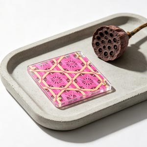 May include: A square coaster featuring a vibrant pink and light yellow geometric star pattern encased in clear acrylic, accompanied by a dried brown lotus seed pod, both resting on a rectangular gray concrete tray with rounded edges.