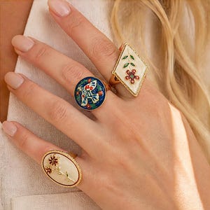 Op de afbeelding: Drie goudkleurige ringen met emaille ontwerpen. Eén ring is ruitvormig met een witte achtergrond en een bloemmotief. Een andere is cirkelvormig met een donkerblauwe achtergrond en een vogelontwerp. De derde is ovaal met een witte achtergrond en bloemen.
