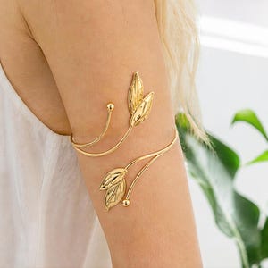 May include: A gold-toned metal arm cuff featuring a wrap-around design with two sets of stylised leaf motifs at the ends. The polished metal band curves around the upper arm, with each leaf element accented by a small spherical bead at the tip.