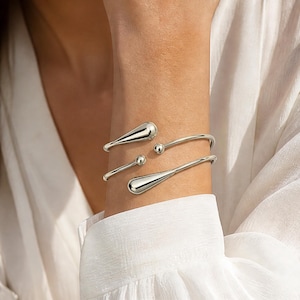 May include: A silver-toned open cuff bracelet featuring a double-wrap design. The ends of the bracelet are finished with two distinct shapes: one side features a smooth, elongated teardrop form, while the other side terminates in a small, polished sphere.
