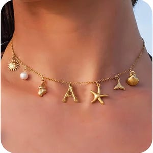 Op de afbeelding: Een goudkleurige ketting met meerdere bedels, waaronder een zon, een witte parel, een schelp, de letter A, een zeester, een vissenstaart en een sint-jakobsschelp. De ketting rust rond de hals en heeft een ontwerp met fijne schakels.