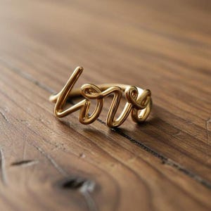 May include: A gold-toned ring featuring the word love written in a continuous cursive script. The metal wire forms the letters l, o, v, and e in a flowing, connected design that wraps around to form the circular band of the ring.