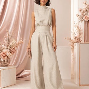 May include: A sleeveless beige jumpsuit featuring a high mock neckline, a fitted waistband, and wide-leg trousers. The garment is crafted from a textured fabric and is paired with a small rectangular clutch and simple black flat sandals.
