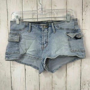 May include: Light blue denim shorts featuring a low-rise waist, a button and zip fly closure, and two side cargo pockets with flap closures. The shorts have a faded wash finish, visible stitching details, and are displayed on a hanger.