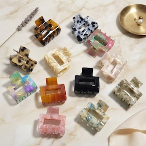 Può includere: Una collezione di dodici mollette rettangolari per capelli in vari motivi e colori, tra cui tartarugato, nero tinta unita, rosa traslucido, bianco marmorizzato, blu e verde, dotate di un meccanismo a molla per fissare i capelli.