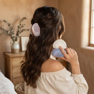 May include: A set of shell-shaped hair clips in pastel colors including light pink, white, and light blue. The clips feature a ribbed, fan-like design with a glossy finish. One clip is secured in dark brown hair, while two others are held.