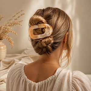 Könnte beinhalten: Eine ovale Haarspange mit einem marmorierten beige-cremefarbenen Muster. Die Spange besteht aus einem glatten, polierten Material und verfügt über ein hohles Design in der Mitte, das eine Haarpartie am Hinterkopf zu einem Dutt zusammenhält.
