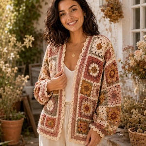 May include: A long-sleeved cardigan made of multi-colored crochet granny squares in shades of dusty rose, mustard yellow, sage green, and cream. The garment features a scalloped cream-colored crochet trim along the front opening and cuffs.