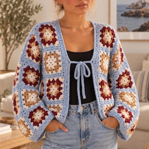 Può includere: Un cardigan all'uncinetto a maniche lunghe con motivo a piastrelle granny nei toni dell'azzurro, crema, beige e bordeaux scuro. Il capo presenta un bordo azzurro, uno scollo a V e una chiusura frontale con laccetti in filato azzurro.