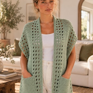 May include: A sage green sleeveless crochet cardigan featuring a vertical openwork grid pattern, a long open front, and two large patch pockets. The garment is layered over a plain white tank top and white trousers, showcasing a textured knit design.