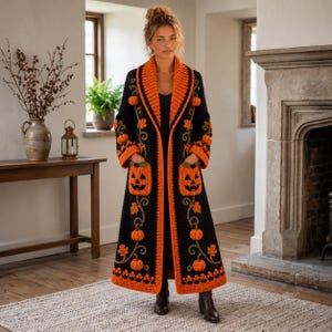 May include: A long, black, open-front knit cardigan featuring a vibrant orange shawl collar and hem. The garment is decorated with crocheted orange pumpkin motifs and green vine patterns, including two large pumpkin-shaped patch pockets.