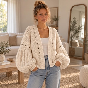 Puede incluir: Un cárdigan de punto color crema abierto por delante, con textura gruesa, escote en pico profundo y mangas abullonadas voluminosas con puños acanalados. La prenda se lleva sobre una camiseta blanca sencilla y unos vaqueros azul claro.
