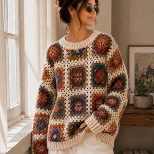 Könnte beinhalten: Ein langärmeliger Pullover aus cremefarbenem Garn mit einem mehrfarbigen Granny-Square-Muster. Das Kleidungsstück verfügt über einen gerippten Rundhalsausschnitt, gerippte Bündchen und einen gerippten Saum in verschiedenen Blau-, Rot- und Brauntönen.