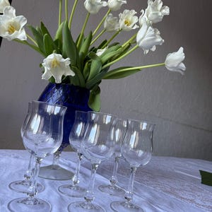 Puede incluir: Un conjunto de cinco copas de cristal transparente con cuencos en forma de tulipán y patrones de líneas horizontales grabadas, dispuestas frente a un jarrón de cristal azul profundo lleno de un ramo de tulipanes blancos.