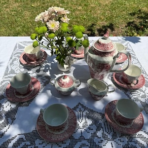Puede incluir: Un juego de té que incluye una tetera, un azucarero, una jarra de leche y cinco tazas con sus platos a juego, decorados con un patrón floral rojo y blanco. Un florero con flores rosas y verdes se encuentra entre las piezas sobre un mantel de encaje.