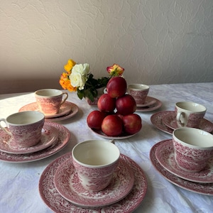 May include: A set of five ceramic teacups and matching saucers featuring a detailed red and white floral pattern. A small bowl holds a cluster of fresh red nectarines, and a small vase contains yellow and white roses on a white tablecloth.