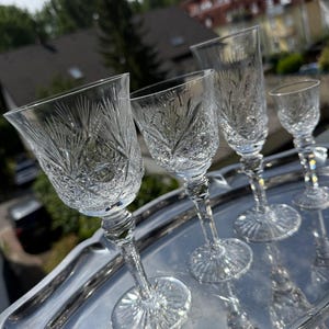 Op de afbeelding: Een set van vier heldere kristallen glazen met ingewikkelde ster- en diamantgeslepen patronen. Elk glas heeft een gefacetteerde steel en een ronde voet met een stralend patroon, geplaatst op een gepolijst zilverkleurig metalen dienblad.