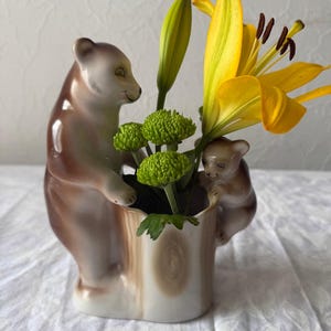 May include: A ceramic vase shaped like a tree stump with two brown bears attached to the sides. The vase holds a bright yellow lily and several small, round, green chrysanthemum flowers, creating a naturalistic display of floral decor.