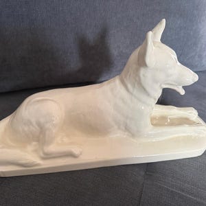May include: A glossy white ceramic figurine of a dog in a lying position. The canine is depicted with pointed ears, a slightly open mouth, and a smooth, reflective surface. The sculpture is mounted on a rectangular base of the same material.