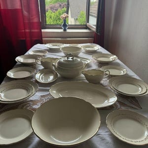 Op de afbeelding: Een grote set crèmekleurig keramieken servies met goudkleurige randen. De set bevat een terrine met deksel, een sauskom, een grote ovale serveerschaal, een grote kom en diverse borden en schaaltjes gerangschikt op een wit tafelkleed.