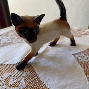 Puede incluir: Una figura de cerámica de un gato siamés con cuerpo de color crema y puntos de color marrón oscuro en la cara, las orejas, la cola y las patas. El gato está representado en posición de pie con la cola levantada, acabado brillante y ojos azules.