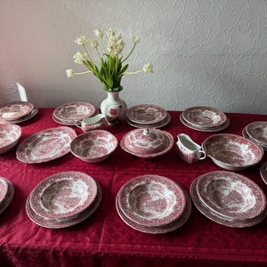 Può includere: Un grande set di stoviglie con un motivo a trasferimento rosso e bianco su piatti, ciotole e piatti da portata in ceramica. La collezione include zuppiere con coperchio, salsiere e ciotole di varie dimensioni, disposte su una tovaglia rossa.