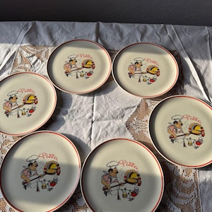 Op de afbeelding: Een set van zes ronde keramische borden met een crèmekleurige basis en een dunne rode rand. Elk bord heeft een centrale illustratie van een kok met een witte muts en rood geruit schort die een pizza in een oven schuift, met het woord Pizza.